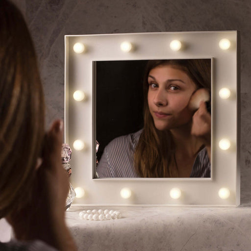 A woman applying makeup in front of a Hollywood Mirror with bright LED lights for flawless beauty routine.