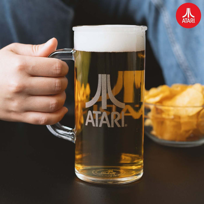 Person holding Official Atari Beer Glass filled with beer, featuring the classic Atari logo, next to a bowl of chips.