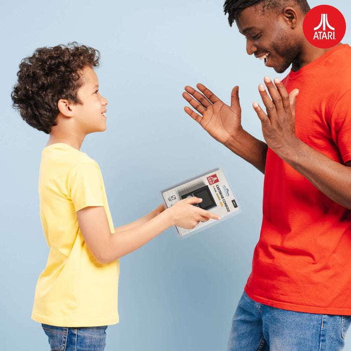 Child in yellow shirt gifting an official Atari handheld console to a man wearing a red shirt against a light blue background.